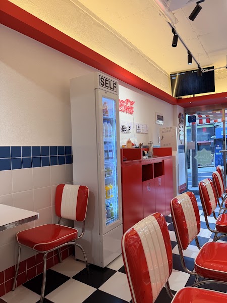 Interior of a restaurant with a checkered floor, red and white diner-style chairs, and a drink refrigerator.