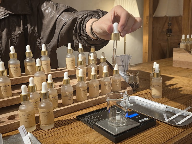 A person holding a glass dropper over a beaker in a perfume studio, surrounded by amber-labeled fragrance bottles on a wooden rack.