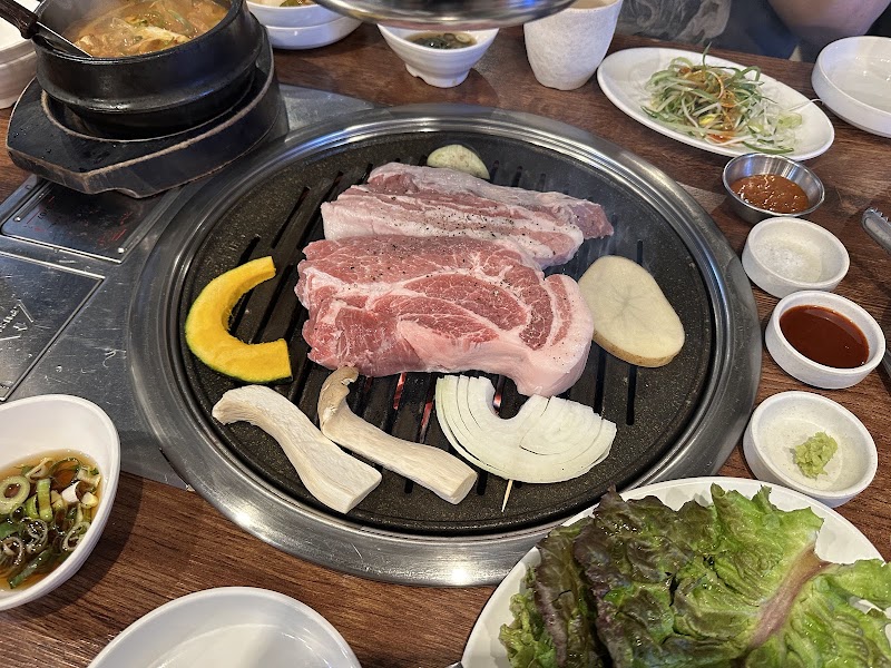A Korean BBQ grill featuring raw pork, pumpkin slices, onions, and mushrooms with various side dishes on a wooden table.