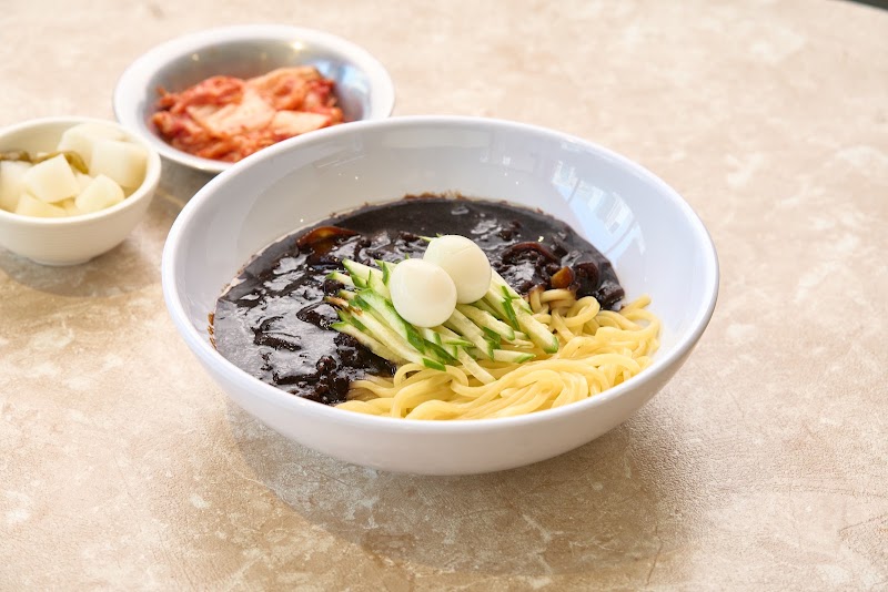 A bowl of Jajangmyeon with black bean sauce, topped with cucumber strips and quail eggs, served with kimchi and pickled radish.
