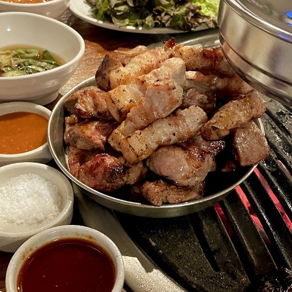 A metal tray of grilled pork belly served at a Korean barbecue restaurant, accompanied by various dipping sauces.