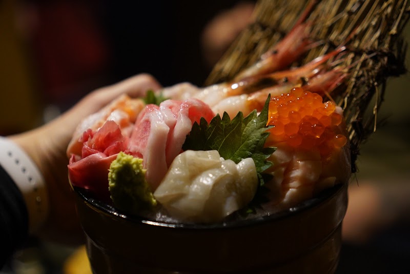 A close-up of a bowl filled with assorted raw fish sashimi, shrimp, and salmon roe held by hand.