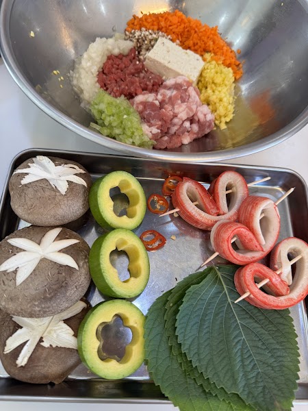 A metal bowl of minced raw ingredients next to a tray of prepared vegetables and garnishes for cooking.