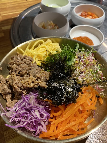 A colorful bibimbap bowl with assorted vegetables, thin beef strips, and seaweed served on a metal table with small side dishes
