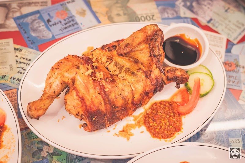 A plate of roasted chicken served with sauce, chili sambal, cucumber, and tomato slices, set against a table covered in paper currency.