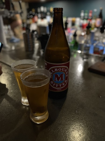 A classic Melbourne Bitter longneck bottle next to glasses on a dark counter