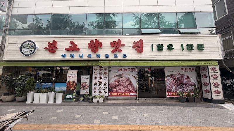 Exterior facade of a Korean BBQ restaurant in Seoul with signage in Korean, large menu posters on glass, and potted plants.
