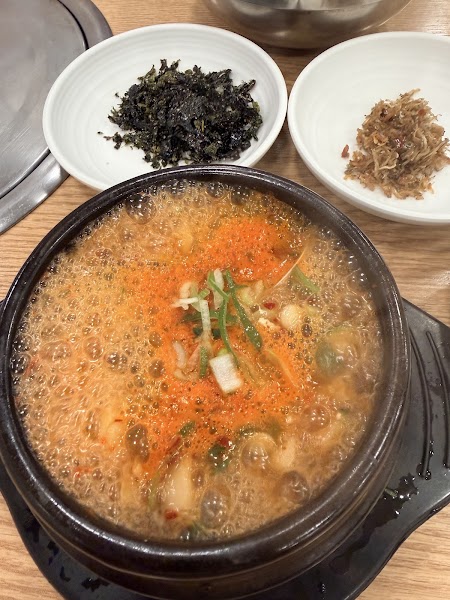 A bubbling hot Korean stew in a black stone pot, served with side dishes of seaweed and dried fish