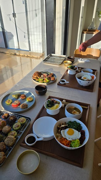 A table set with multiple bowls of Korean bibimbap, side dishes, and small plates of pan-fried vegetables and patties.