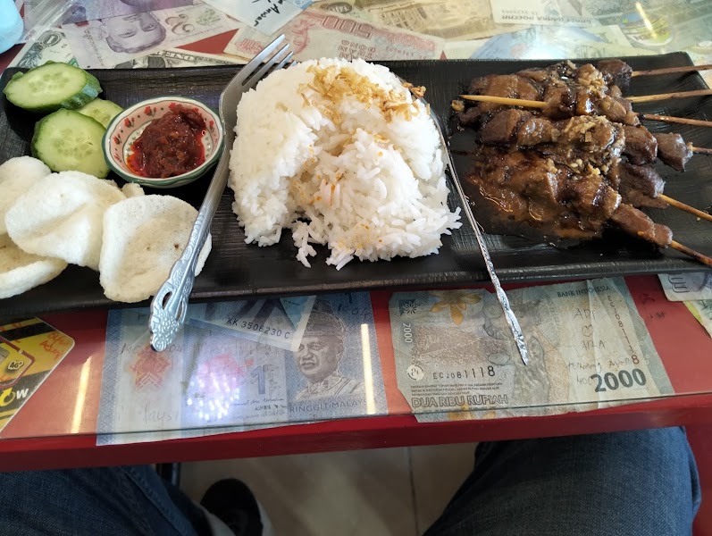 A serving of chicken satay with rice, cucumber slices, chili paste, and crackers served on a black platter over a table covered in banknotes.