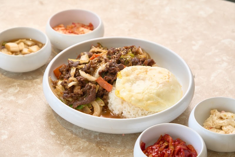 A white bowl containing beef stir-fry with rice and a fried egg, surrounded by four small side dishes on a marble-patterned table.