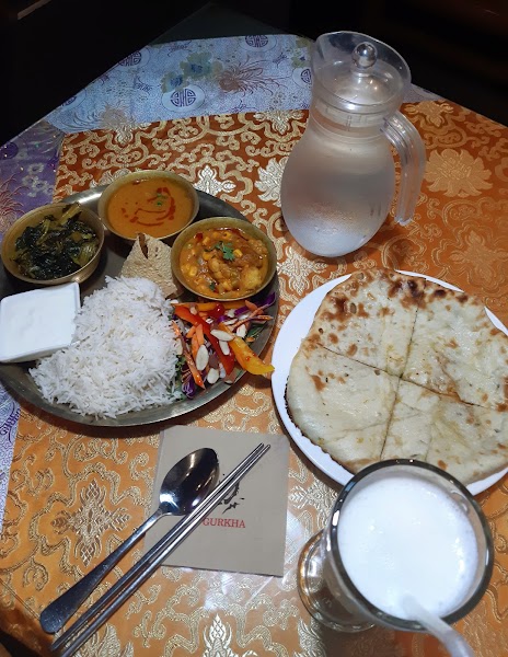 An Indian thali platter with rice, curry, naan, a water pitcher, and a lassi drink on a patterned tablecloth