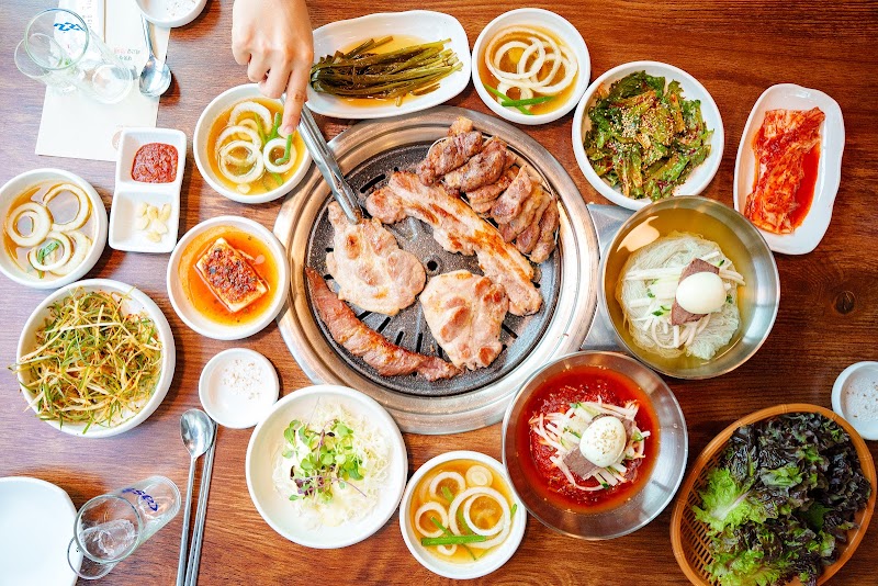 Top-down view of a Korean barbecue feast with grilled meat, various side dishes, and cold noodles on a wooden table.
