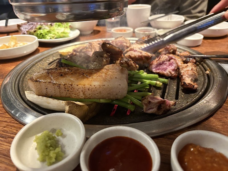 Close-up of thick pork belly and green vegetables grilling on a metal circular grill at a Korean barbecue restaurant.