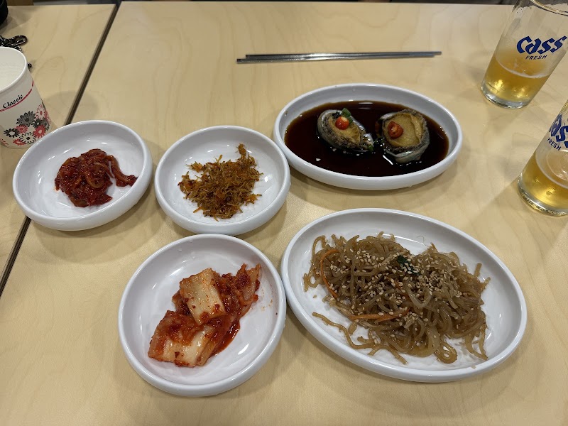 A spread of various Korean side dishes, including kimchi, abalone, and glass noodles, on a light-colored restaurant table