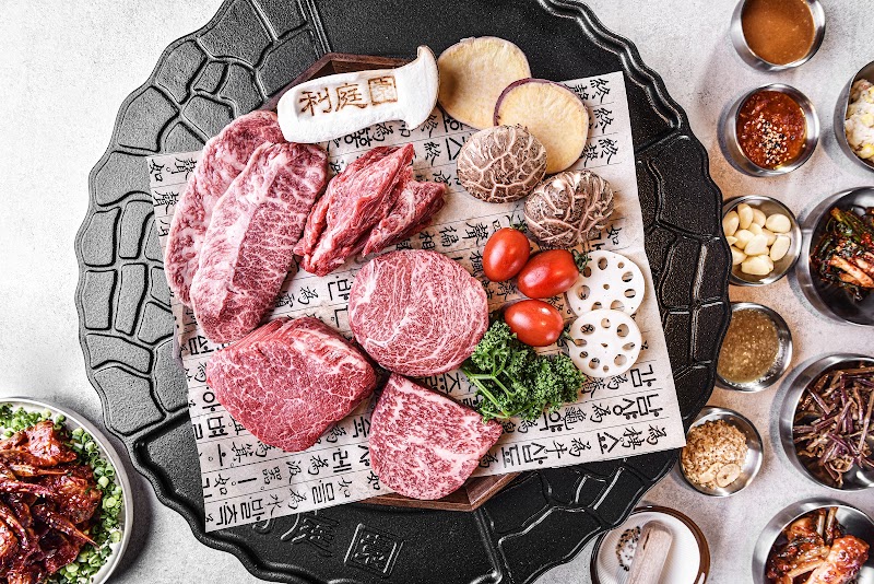 A flat-lay of raw marbled beef cuts, mushrooms, and various Korean side dishes served on a dark textured platter.