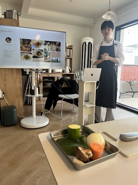 Chef in an apron standing in a kitchen with a screen presentation and fresh vegetables on a tray in the foreground.
