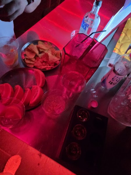 A high-angle view of a bar table with chips, snacks, and a vodka bottle under vibrant red neon lighting