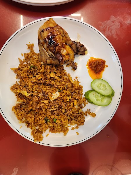 A plate of fried rice with chicken, chili sauce, and cucumber slices on a red table.