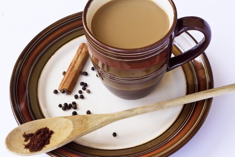 A cup of creamy milk tea on a saucer with a cinnamon stick, peppercorns, and a wooden spoon with spice powder