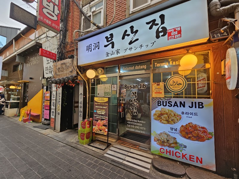 Exterior view of Busan Jib restaurant in Myeongdong, featuring a brightly lit entrance, signage, and a chicken menu advertisement