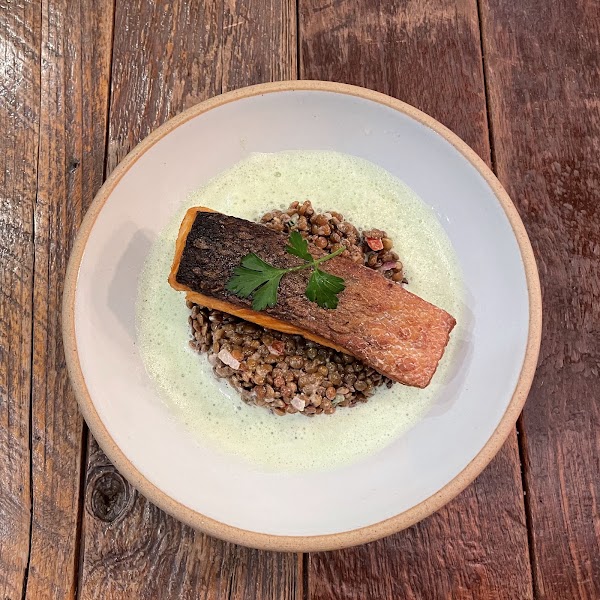 A pan-seared salmon fillet over a bed of lentils with green herb foam in a white bowl on a rustic wood table.