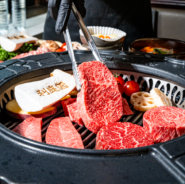 A chef using tongs to place raw marbled beef onto a Korean barbecue grill alongside vegetables.