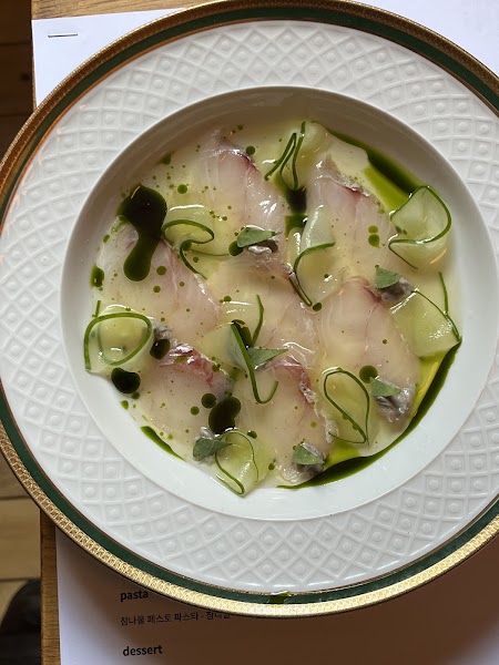 Decorative plate of thinly sliced translucent fish garnished with green herb oil and cucumber ribbons