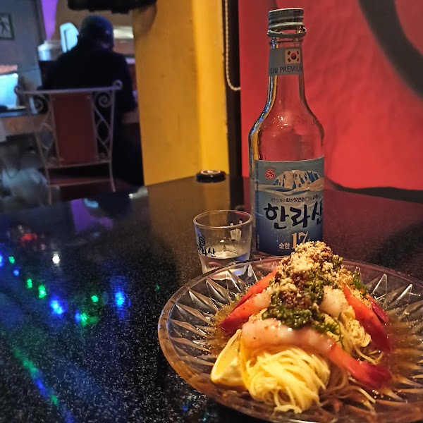 A plate of shrimp noodle dish and a bottle of Korean soju on a black table inside a dimly lit bar.