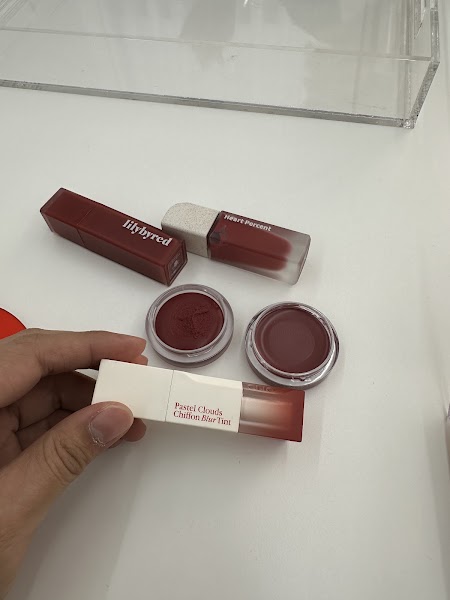 A hand holding a lipstick container with open jars of cream makeup on a white surface.