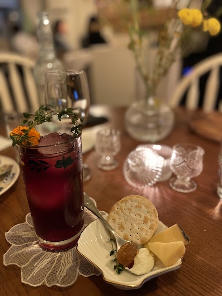 Red artisanal cocktail garnished with fresh herbs and orange flower beside cheese plate