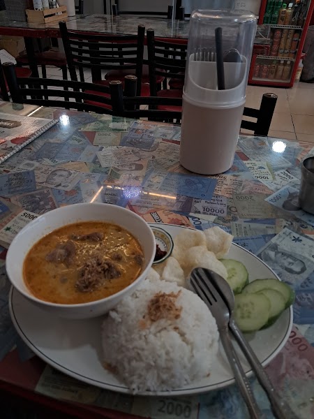 A plate of Indonesian-style curry, rice, crackers, and cucumber slices served on a table covered with Indonesian Rupiah banknotes.