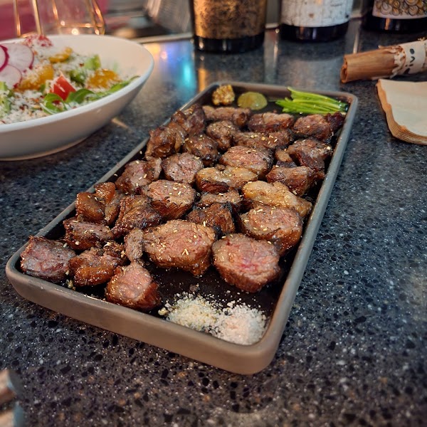 A rectangular plate of grilled steak cubes served with mustard, chives, and salt on a dark table