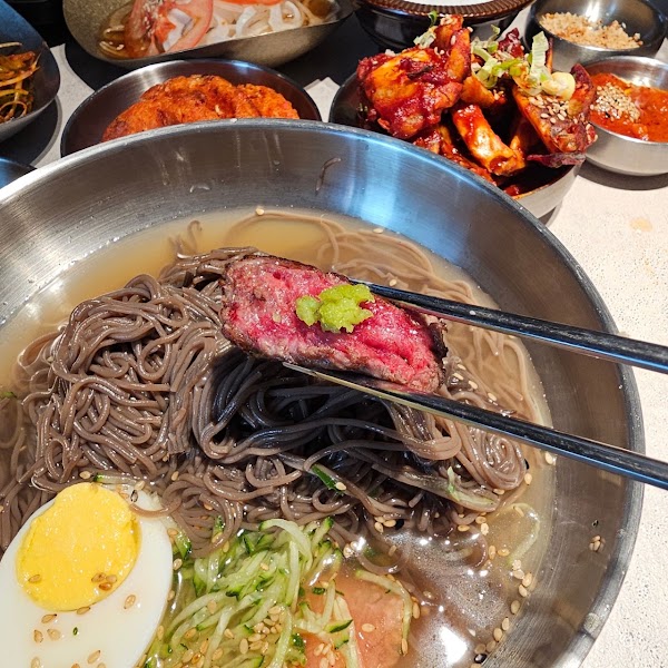 Close-up of metal chopsticks holding a slice of rare seared beef with wasabi over a bowl of cold buckwheat noodles in broth.