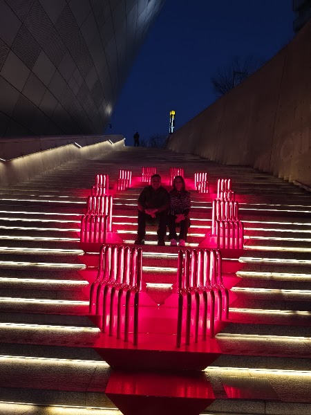 Two people sitting on stairs illuminated by bright red light art installation at Dongdaemun Design Plaza at night