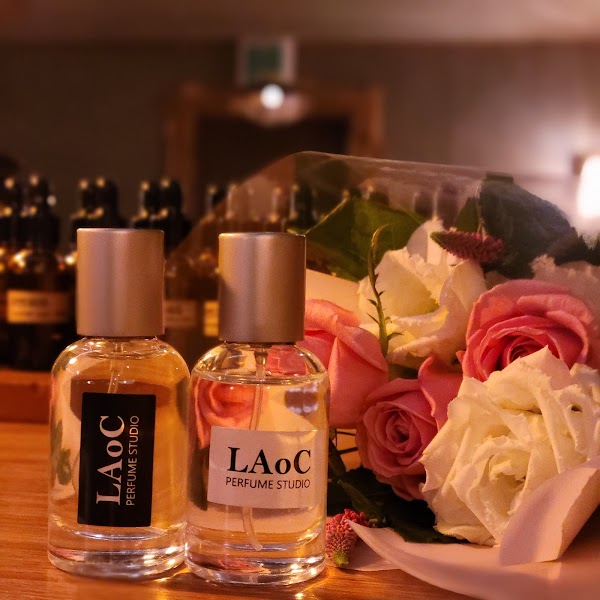 Two clear glass perfume bottles labeled LAoC Perfume Studio sitting next to a bouquet of pink and white roses.