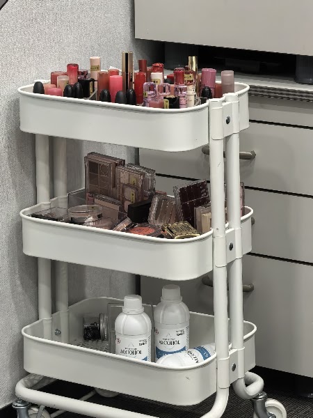 Three-tiered white utility cart filled with makeup palettes, lipsticks, and diagnostic tools