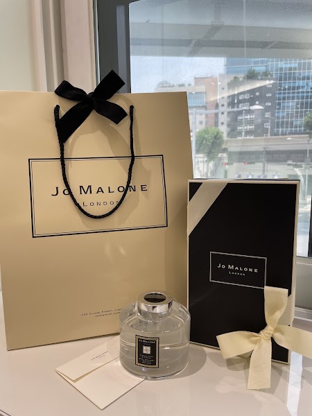 Jo Malone shopping bag, gift box, and scent diffuser with a small card on a white surface by a window