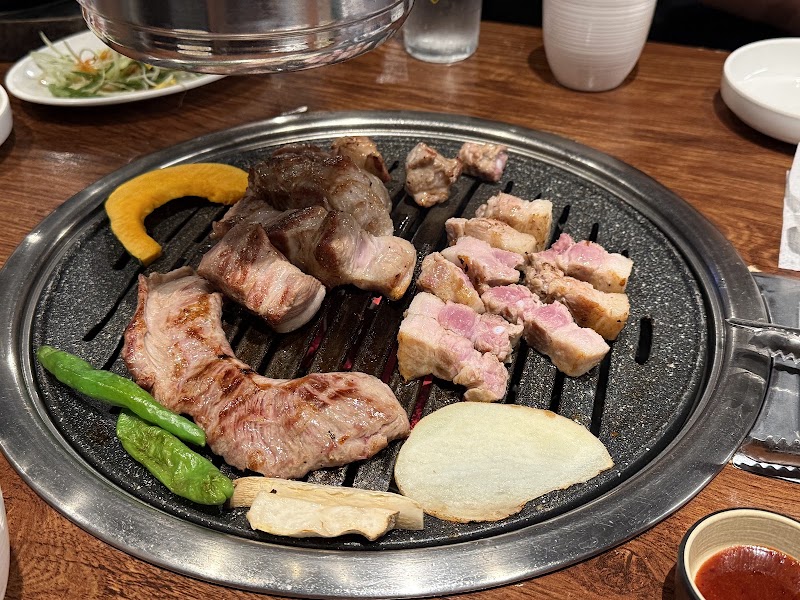 Slabs of pork belly, pumpkin, and green peppers grilling on a circular Korean BBQ grill at a restaurant table.