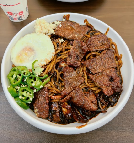 A white bowl containing noodles with dark sauce, topped with seared meat slices, a fried egg, chopped green chili, and minced garlic.
