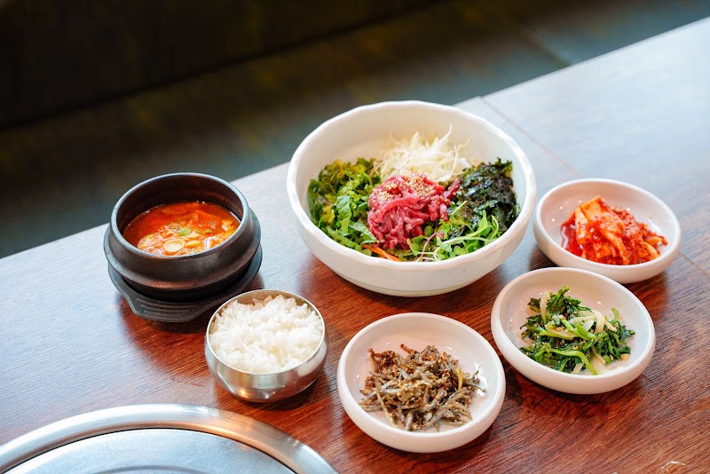 A Korean meal set with a raw beef bibimbap bowl, spicy soup, rice, and various side dishes on a wooden restaurant table.
