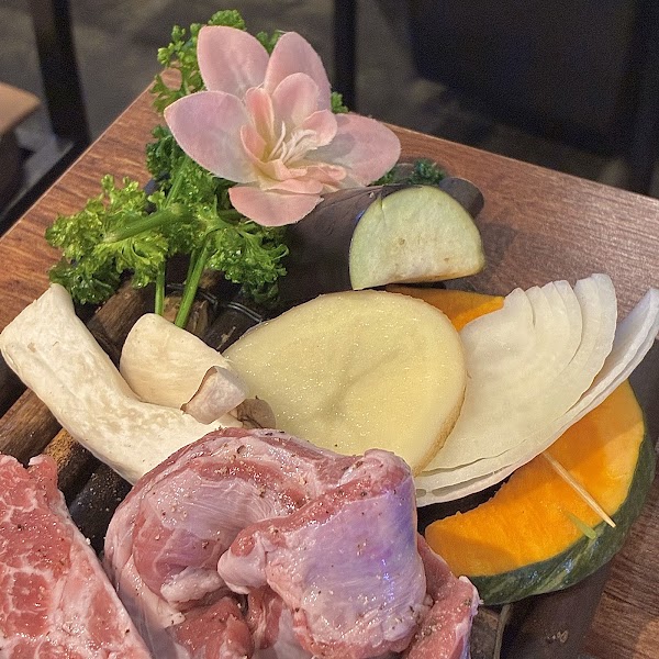 A wooden tray holding raw pork, slices of potato, pumpkin, eggplant, mushroom, onion, and a decorative pink flower.