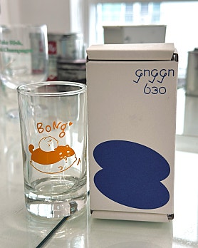 A clear glass with a cute orange animal graphic standing next to a white packaging box with blue abstract design.
