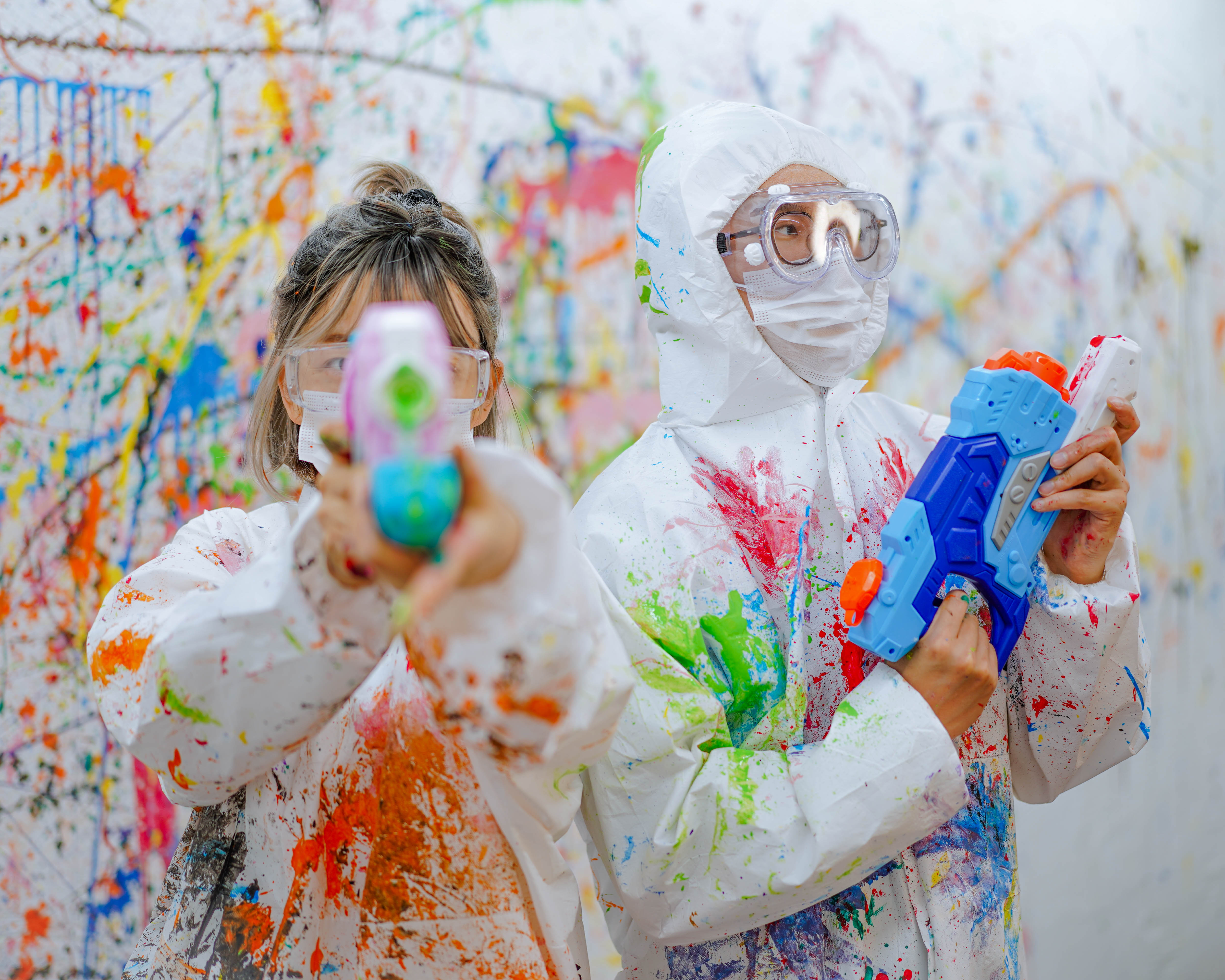 Two people in white protective coveralls, goggles, and masks playing with water guns in a paint-splattered studio