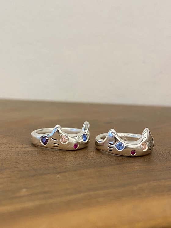 Two cat-shaped silver rings with embedded colorful gemstones resting on a wooden surface