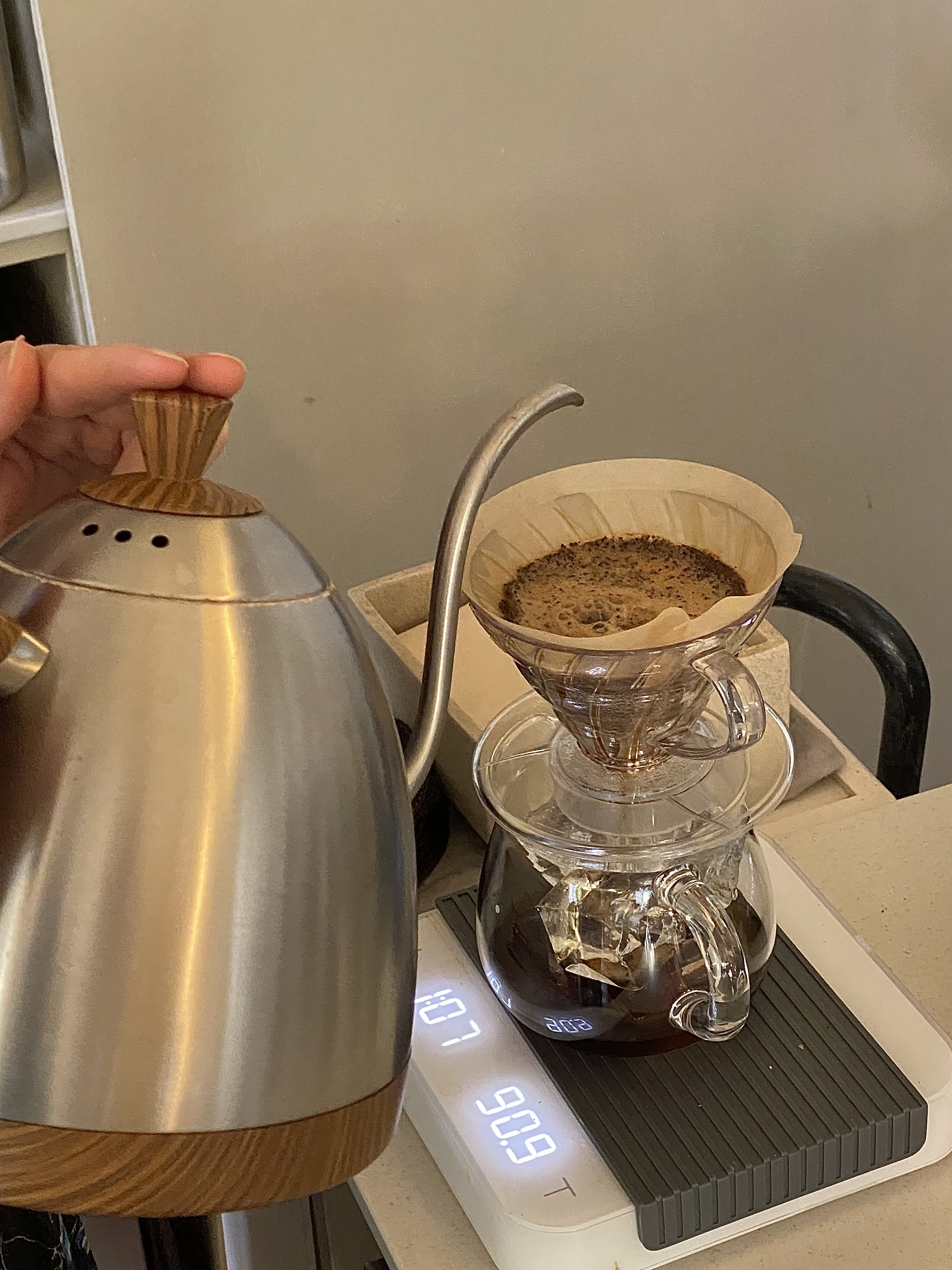 Barista pouring hot water from a gooseneck kettle into a glass coffee dripper over a digital scale