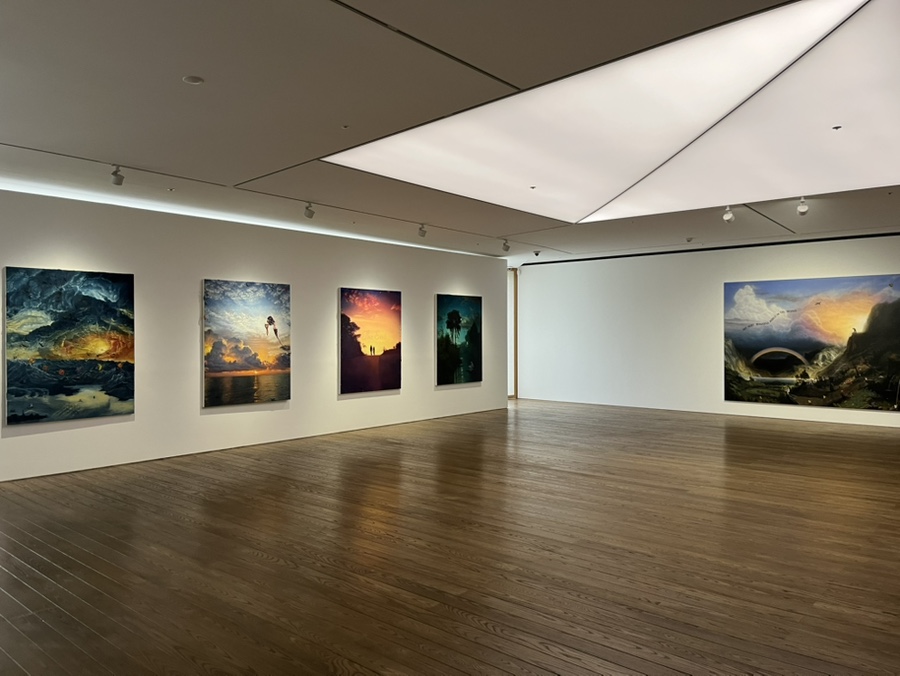 An art gallery interior with wooden flooring and white walls displaying several landscape paintings.