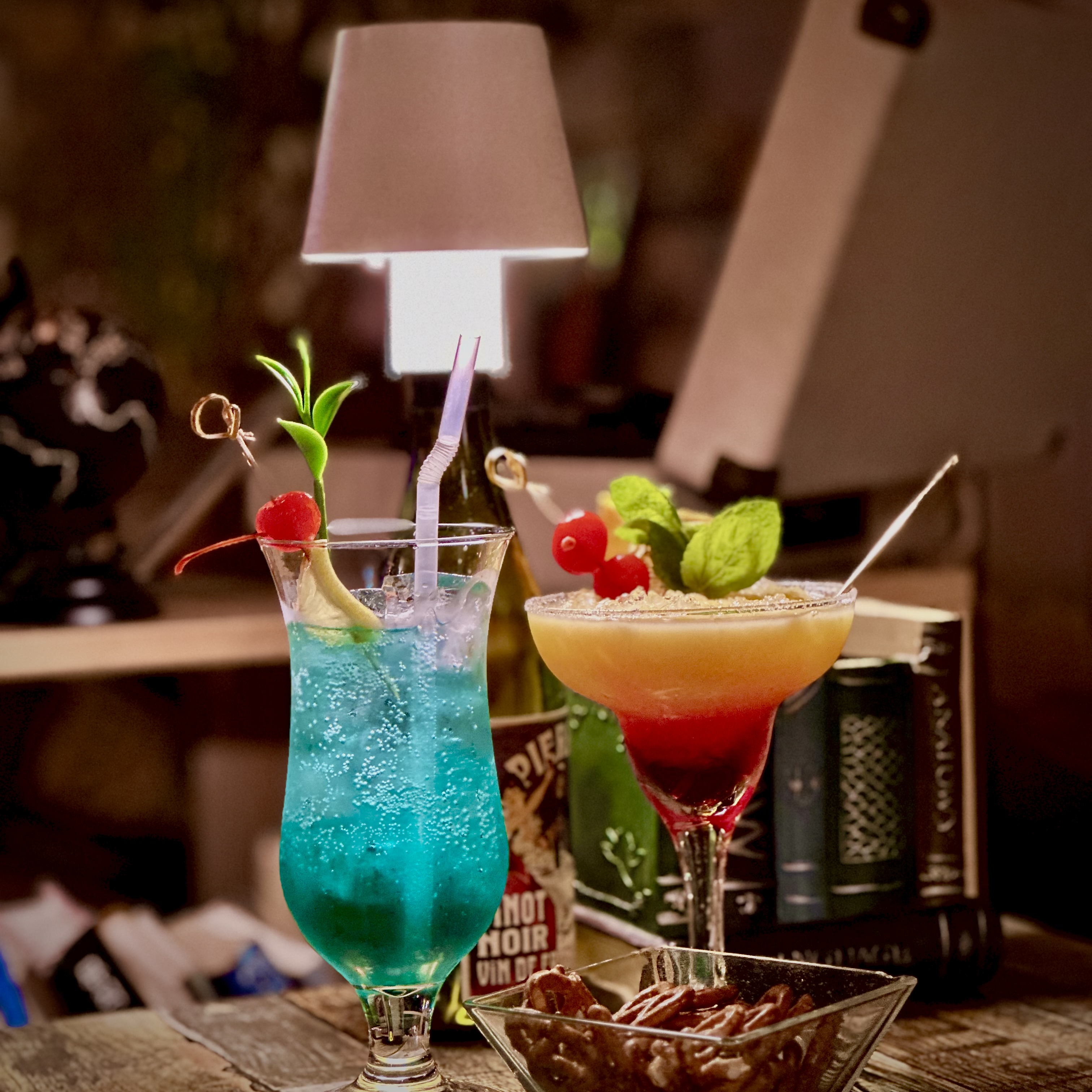 Two colorful cocktails on a table with a snack bowl and small table lamp