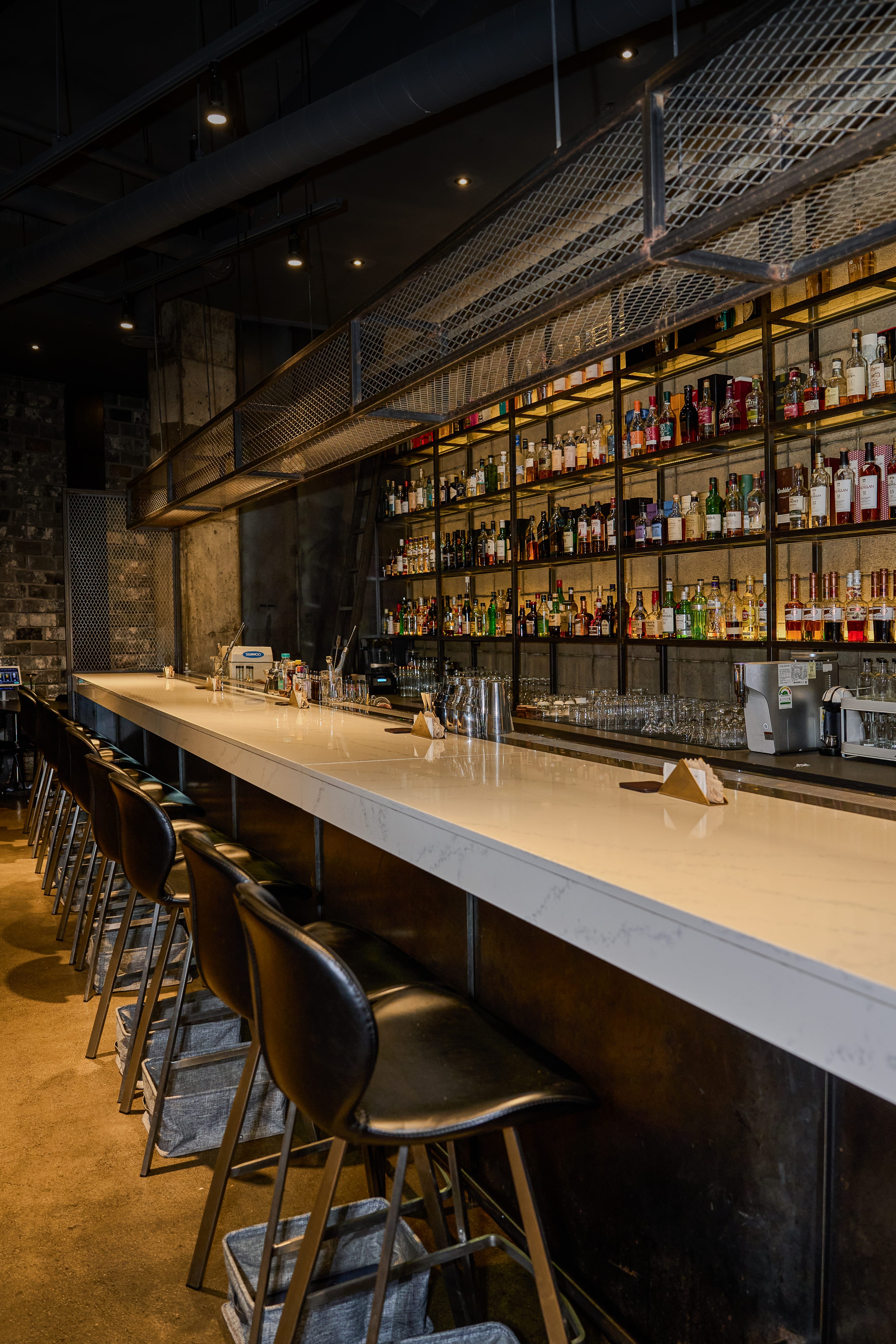 Interior of Bitters bar in Seoul featuring a long marble-top counter, black bar stools, and shelves filled with liquor bottles.