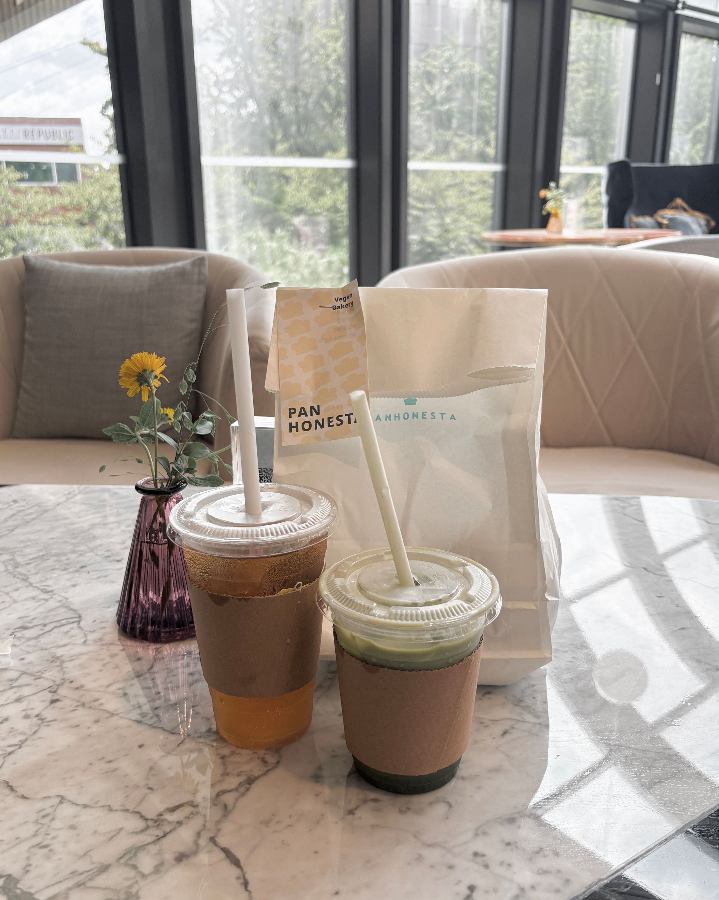 Two plastic cups with drinks and a branded paper bag on a marble table in a sunny cafe.